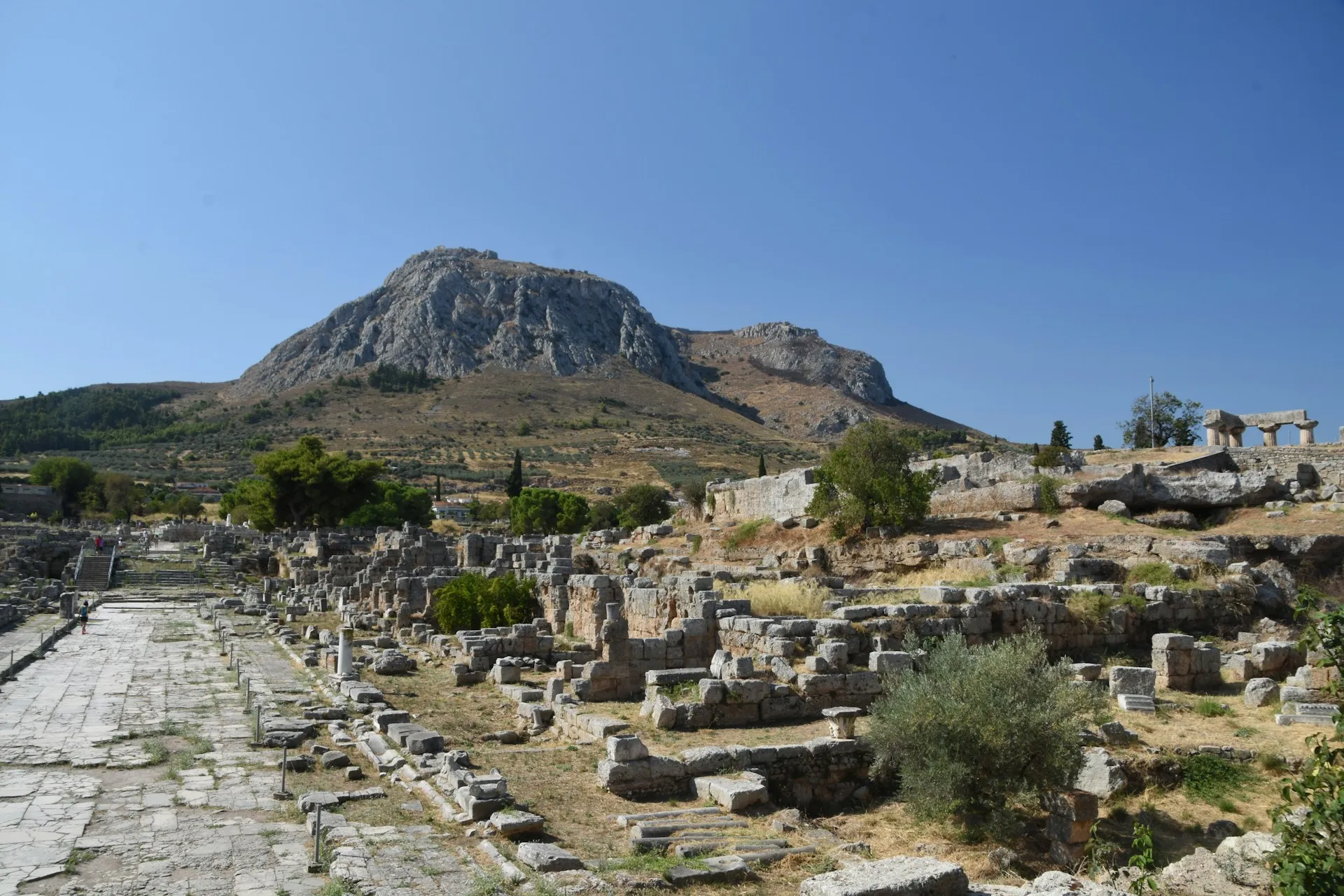 Ruins of Ancient Corinth with a long stone walkway and Acrocorinth rising in the background.