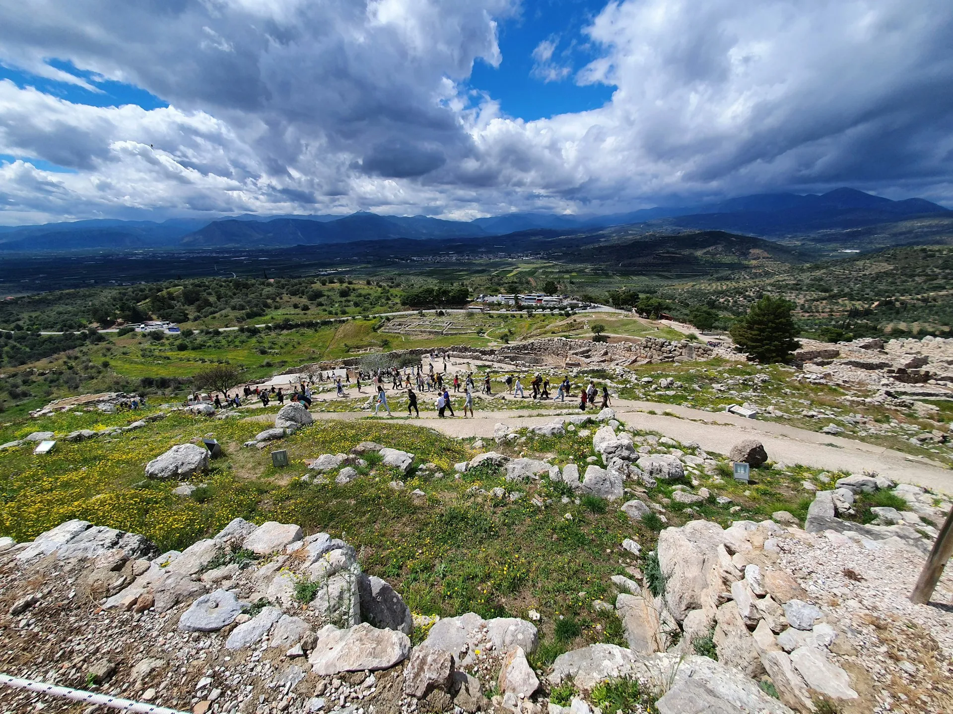 Visitors walking among the ruins of Mycenae with a broad mountain valley under dramatic clouds.